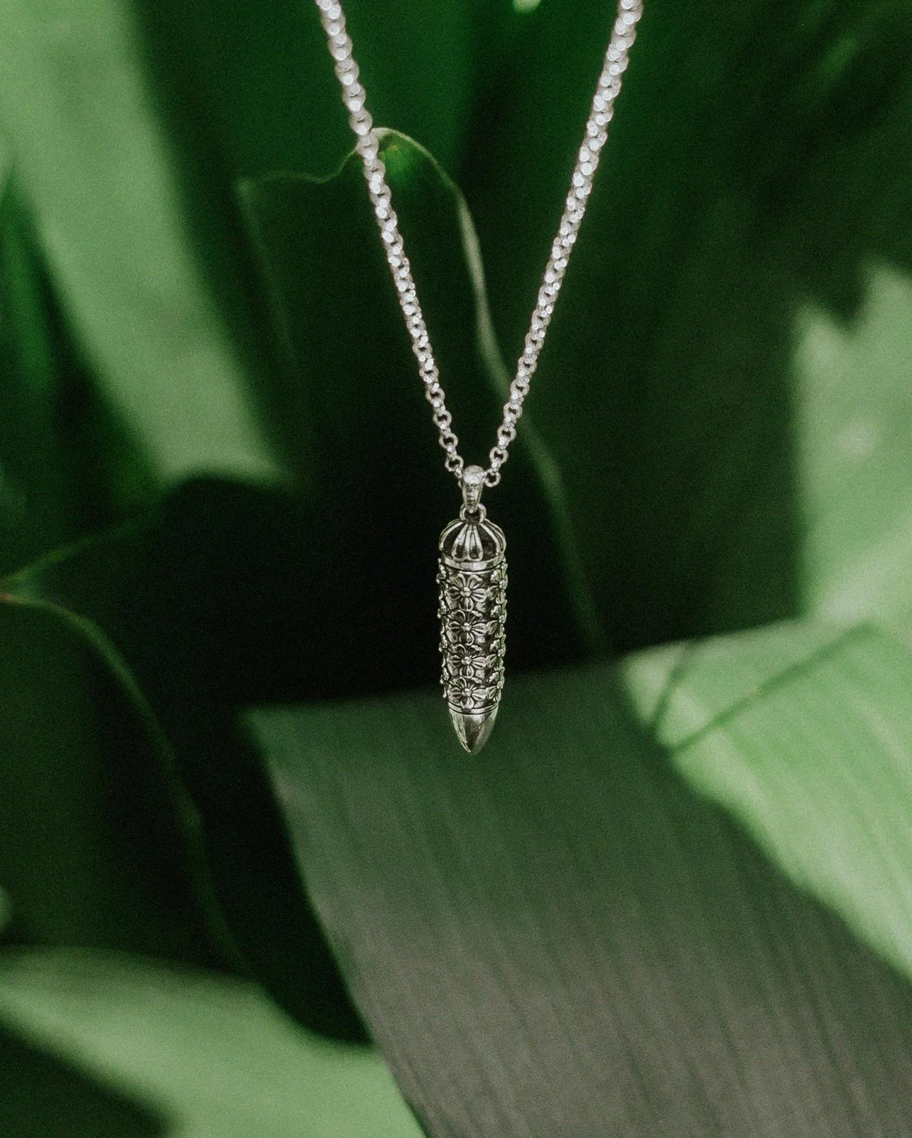 Bullet Necklace with a silver flower-covered pendant against green leaves.